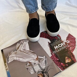 May include: A pair of black suede platform slip-on shoes with white soles. The shoes are being worn by a person wearing blue jeans.