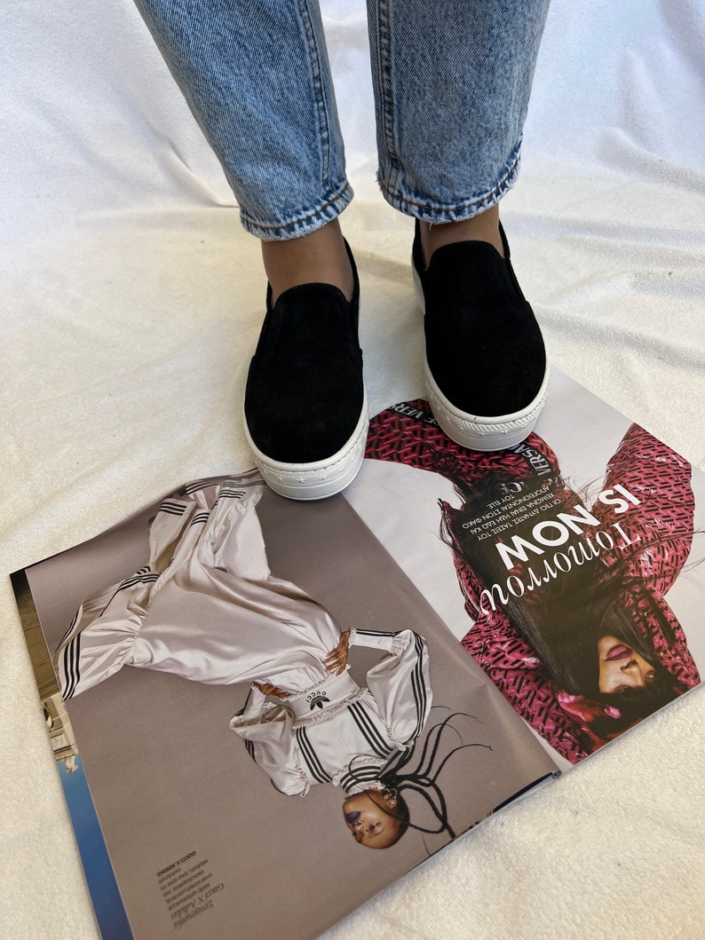 May include: A pair of black suede platform slip-on shoes with white soles. The shoes are being worn by a person wearing blue jeans.