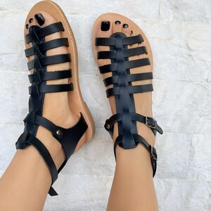 May include: A pair of black gladiator sandals featuring multiple horizontal straps across the foot, a vertical center strap, and an adjustable ankle strap with a silver-toned metal buckle. The sandals have a flat, tan-colored sole.