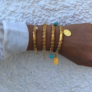 Pode incluir: Cinco pulseiras de ouro com designs decorativos e pingentes. Os pingentes incluem uma p&eacute;rola, uma conta turquesa e um pingente de ouro com uma cruz.