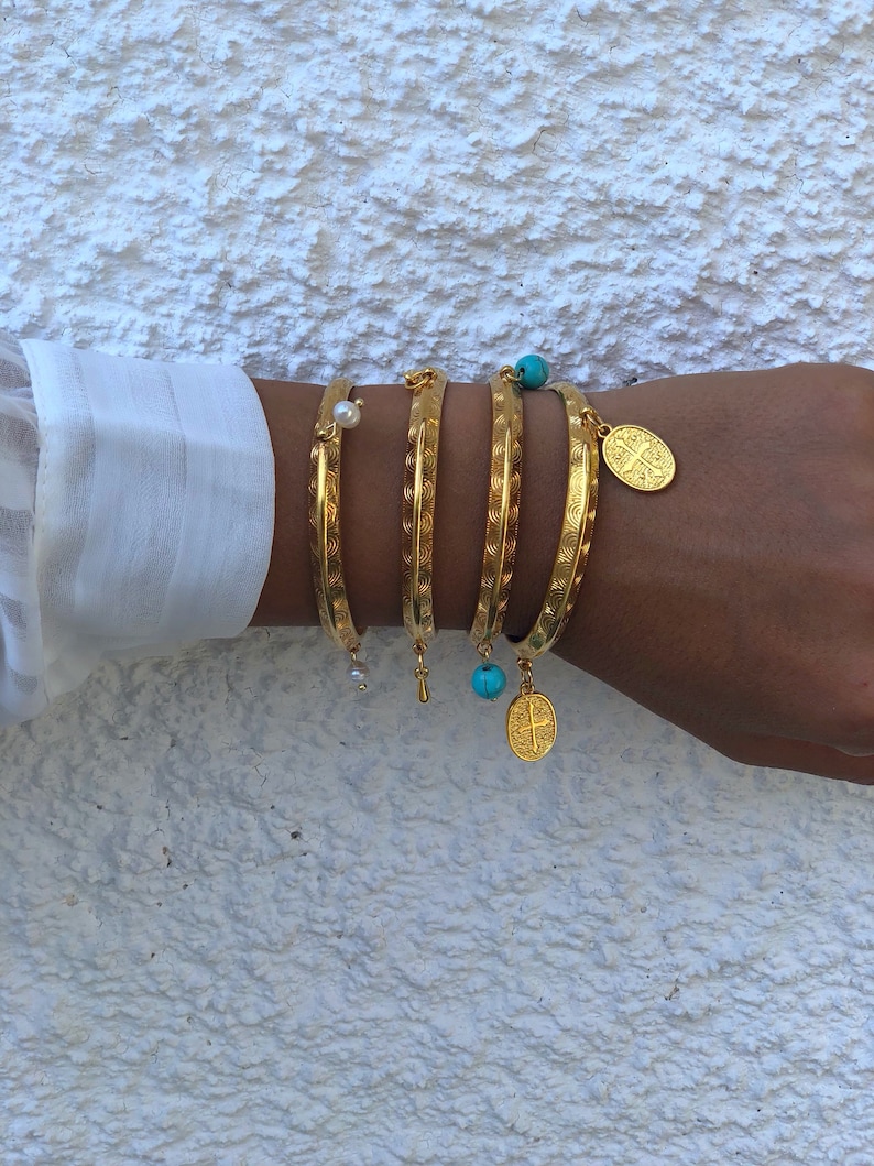 Pode incluir: Cinco pulseiras de ouro com designs decorativos e pingentes. Os pingentes incluem uma p&eacute;rola, uma conta turquesa e um pingente de ouro com uma cruz.
