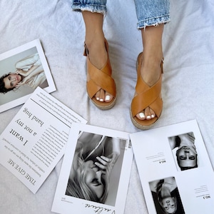 May include: Tan leather sandals with a woven platform sole. The sandals have a criss-cross strap design and a buckle closure. The image also shows pages from a magazine with black and white photos and the text "Vulture."
