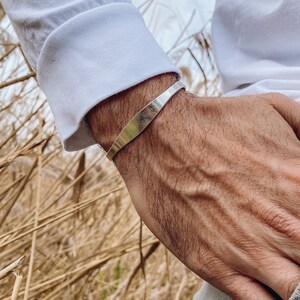 May include: A silver cuff bracelet with a slightly curved design. The bracelet is worn on a person's wrist.