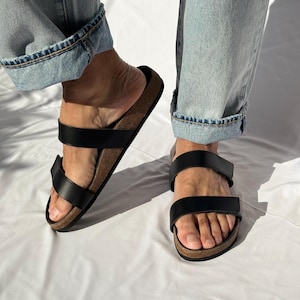 May include: Black leather sandals with two adjustable straps and a cork footbed. The sandals are worn with light blue denim jeans. The sandals are a casual footwear choice.