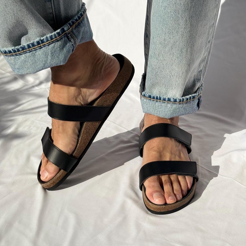 May include: Black leather sandals with two adjustable straps and a cork footbed. The sandals are worn with light blue denim jeans. The sandals are a casual footwear choice.