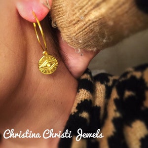 Puede incluir: Un pendiente de aro dorado con un dije circular con un dise&ntilde;o de ojo. El pendiente se lleva en la oreja. La imagen tambi&eacute;n muestra un peque&ntilde;o pendiente de bot&oacute;n plateado y el texto "Christina Christi Jewels".