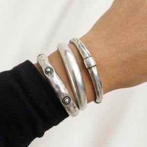 Può includere: Vengono mostrati quattro braccialetti d'argento di vari design. Un braccialetto ha una trama martellata con accenti circolari decorativi. Un altro è liscio e arrotondato, mentre il terzo ha una superficie strutturata con una chiusura. I braccialetti sono indossati su un polso.
