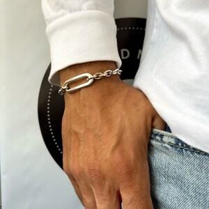 K&ouml;nnte beinhalten: Ein silbernes Kettenarmband mit einem gro&szlig;en ovalen Glied. Das Armband befindet sich am Handgelenk einer Person.