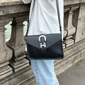 Puede incluir: Un bolso bandolera de cuero negro con una hebilla plateada y tachuelas. El bolso tiene cierre de solapa y una correa larga ajustable.