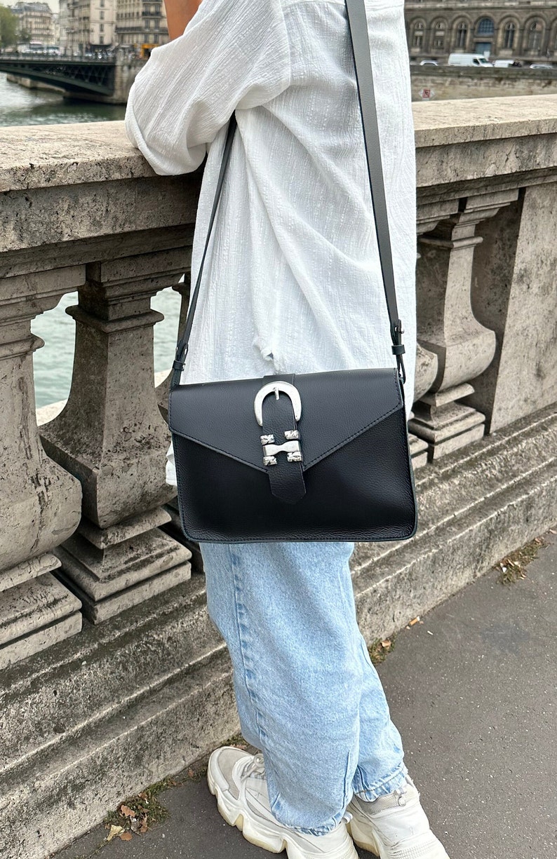Puede incluir: Un bolso bandolera de cuero negro con una hebilla plateada y tachuelas. El bolso tiene cierre de solapa y una correa larga ajustable.