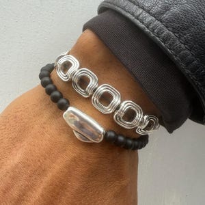 May include: Two bracelets. The top bracelet is silver with a series of square-shaped links. The bottom bracelet is black beaded with a silver oval-shaped accent. The bracelets are worn on a wrist.