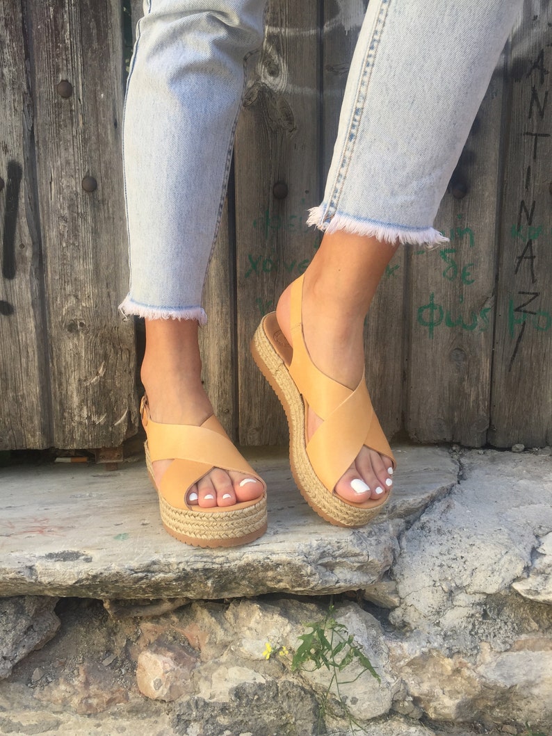 May include: Tan leather platform sandals with crisscross straps and a woven jute sole. The sandals have a slingback strap and are worn with light wash denim jeans with frayed hems. The sandals are approximately 2 inches high.