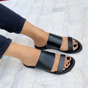 May include: A pair of black leather sandals with two straps across the foot. The sandals have a flat sole and are open-toed.