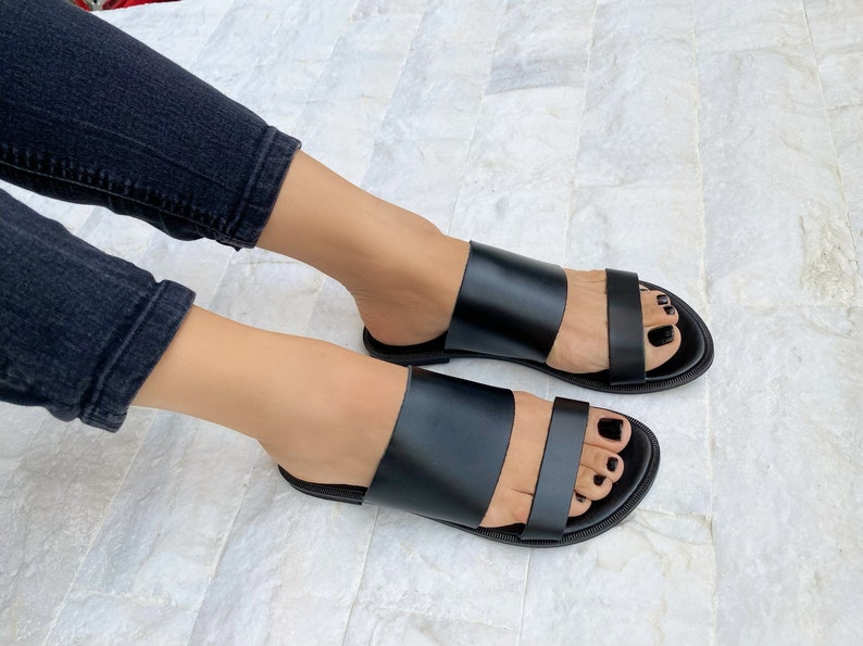 May include: A pair of black leather sandals with two straps across the foot. The sandals have a flat sole and are open-toed.
