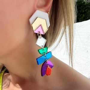 Op de afbeelding: Geometrische oorhangers met een wit, goud en roze bovendeel, gevolgd door een reeks kleurrijke vormen in groen, blauw, paars en rood. De oorbellen zijn bevestigd met goudkleurige ringen en zijn ontworpen om op te vallen.
