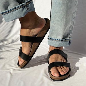 May include: Black leather sandals with two adjustable straps and a cork footbed. The sandals are worn with light blue denim jeans. The sandals are a comfortable and stylish footwear option.