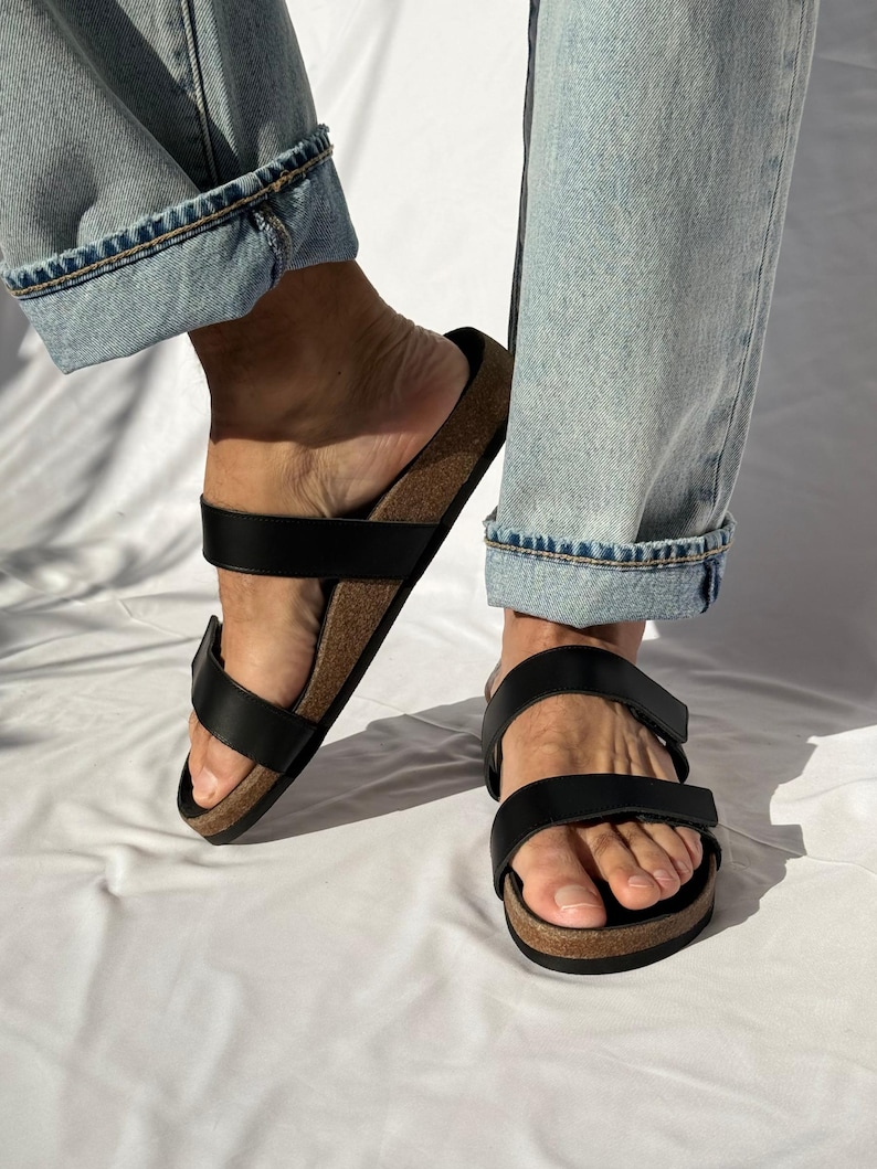 May include: Black leather sandals with two adjustable straps and a cork footbed. The sandals are worn with light blue denim jeans. The sandals are a comfortable and stylish footwear option.