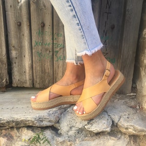 May include: Tan leather platform sandals with crisscross straps and a buckle closure. The sandals have a woven jute platform sole. The person is wearing light blue jeans with frayed hems.