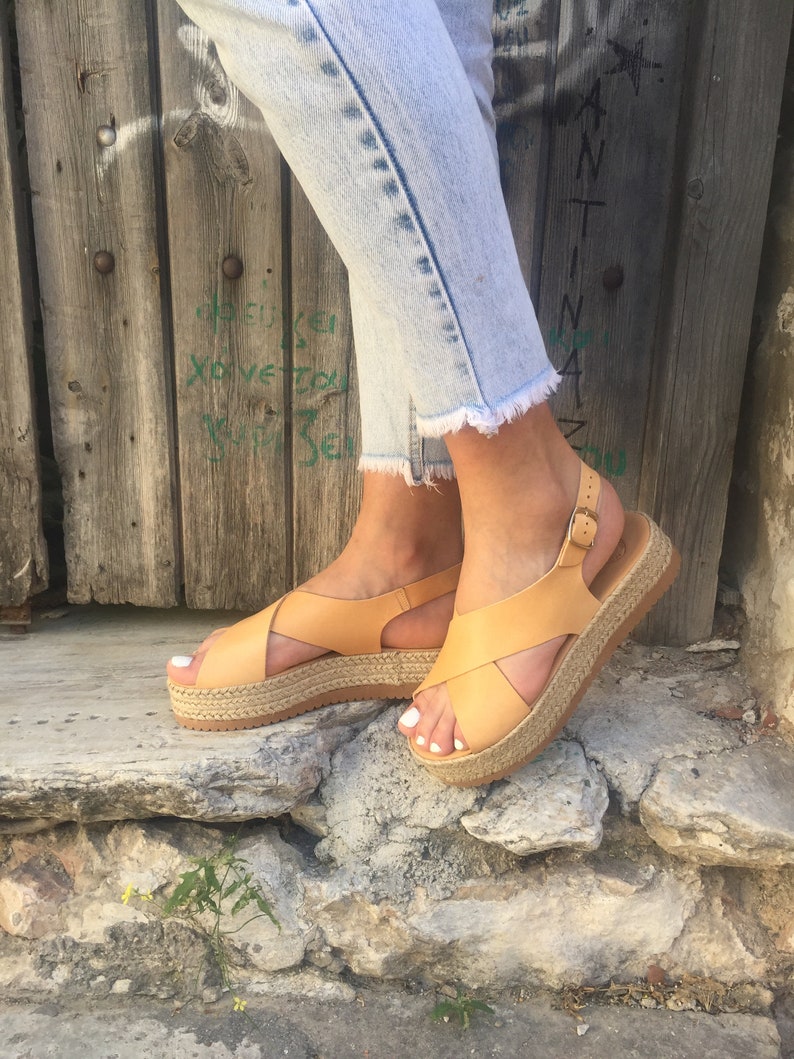 May include: Tan leather platform sandals with crisscross straps and a buckle closure. The sandals have a woven jute platform sole. The person is wearing light blue jeans with frayed hems.
