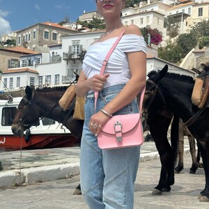 Op de afbeelding: Een vrouw draagt een wit off-shoulder topje, blauwe jeans en een zonnebril en draagt een roze leren schoudertas met een zilveren gesp. Ze staat voor een wit gebouw met een ezel op de achtergrond.