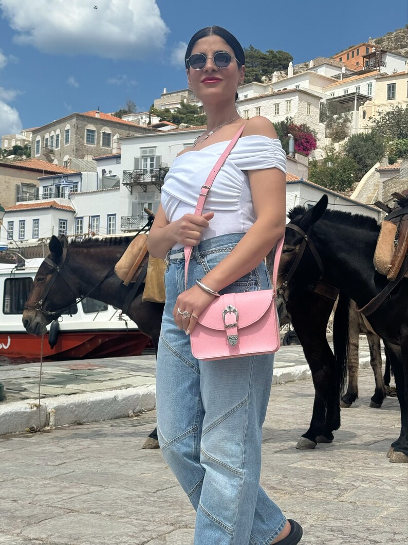 Op de afbeelding: Een vrouw draagt een wit off-shoulder topje, blauwe jeans en een zonnebril en draagt een roze leren schoudertas met een zilveren gesp. Ze staat voor een wit gebouw met een ezel op de achtergrond.