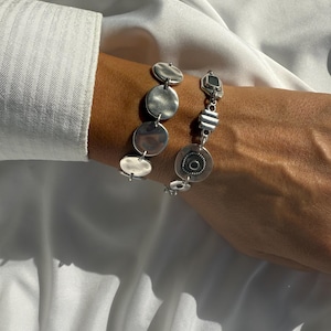 May include: Two silver bracelets are displayed on a person's wrist. One bracelet features a series of connected, flat, circular charms. The other bracelet has a mix of geometric shapes and a circular charm with a central design. The bracelets are set against a white background.