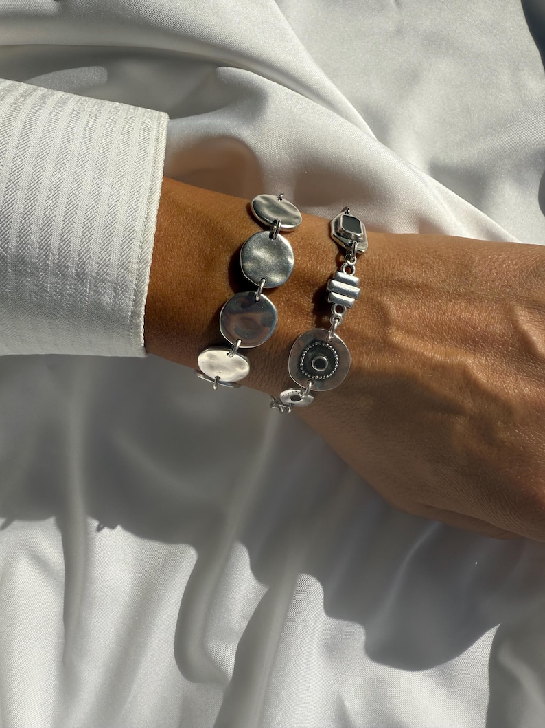 May include: Two silver bracelets are displayed on a person's wrist. One bracelet features a series of connected, flat, circular charms. The other bracelet has a mix of geometric shapes and a circular charm with a central design. The bracelets are set against a white background.