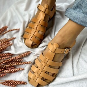 May include: A pair of tan leather sandals with a caged design. The sandals have a buckle closure and are shown on a white fabric background.