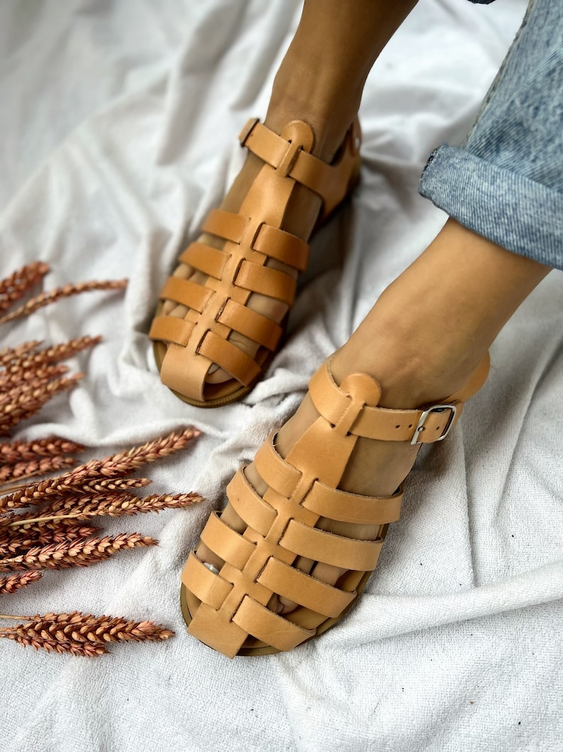 May include: A pair of tan leather sandals with a caged design. The sandals have a buckle closure and are shown on a white fabric background.