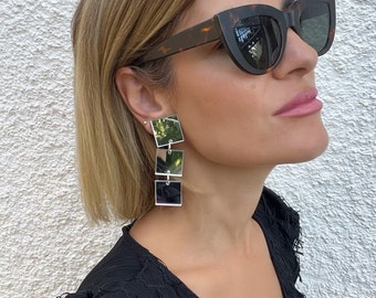 May include: A woman wearing tortoiseshell cat-eye sunglasses and silver mirrored square earrings. The earrings are layered and dangle from the earlobe.