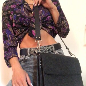 Puede incluir: Un bolso bandolera de cuero negro con cierre de solapa y hebilla plateada. El bolso tiene una superficie texturizada y una correa larga ajustable.