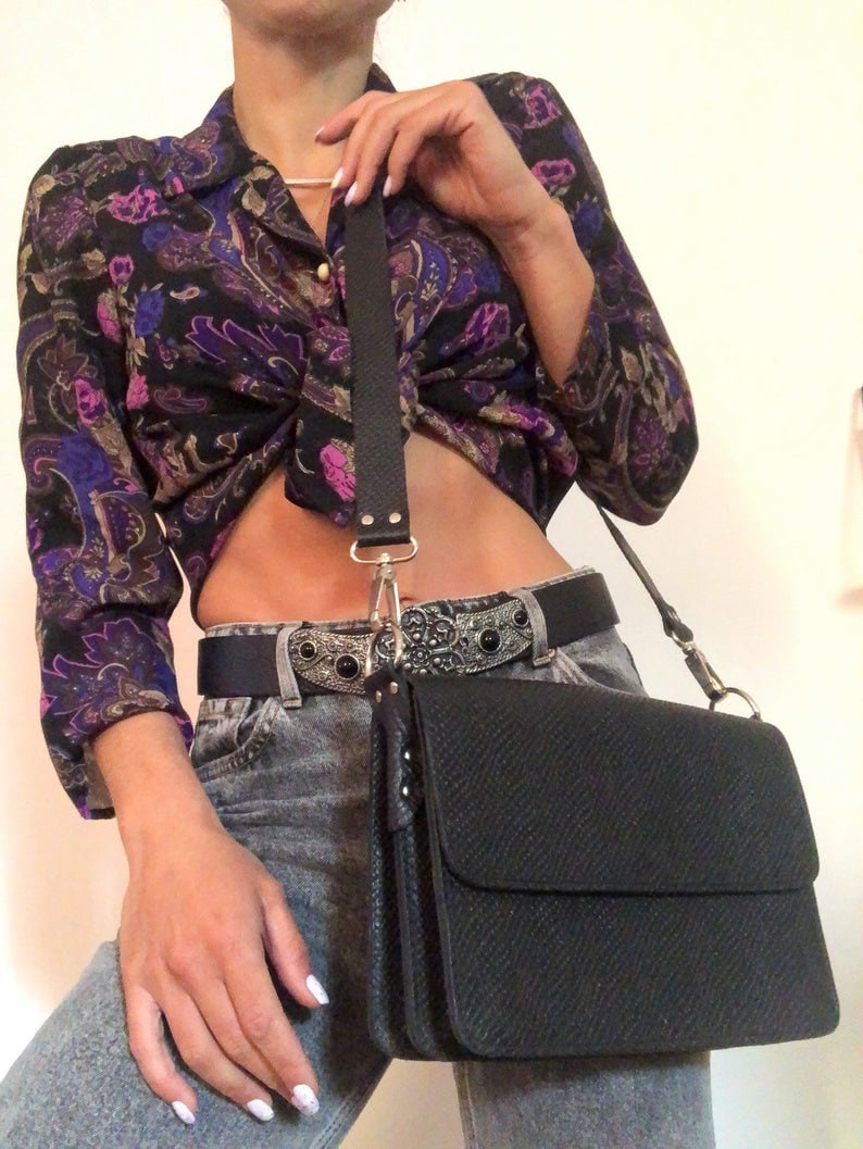 Puede incluir: Un bolso bandolera de cuero negro con cierre de solapa y hebilla plateada. El bolso tiene una superficie texturizada y una correa larga ajustable.