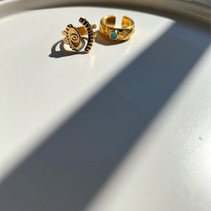 May include: Two gold rings on a white surface. One ring features an eye design and the other has a turquoise stone.