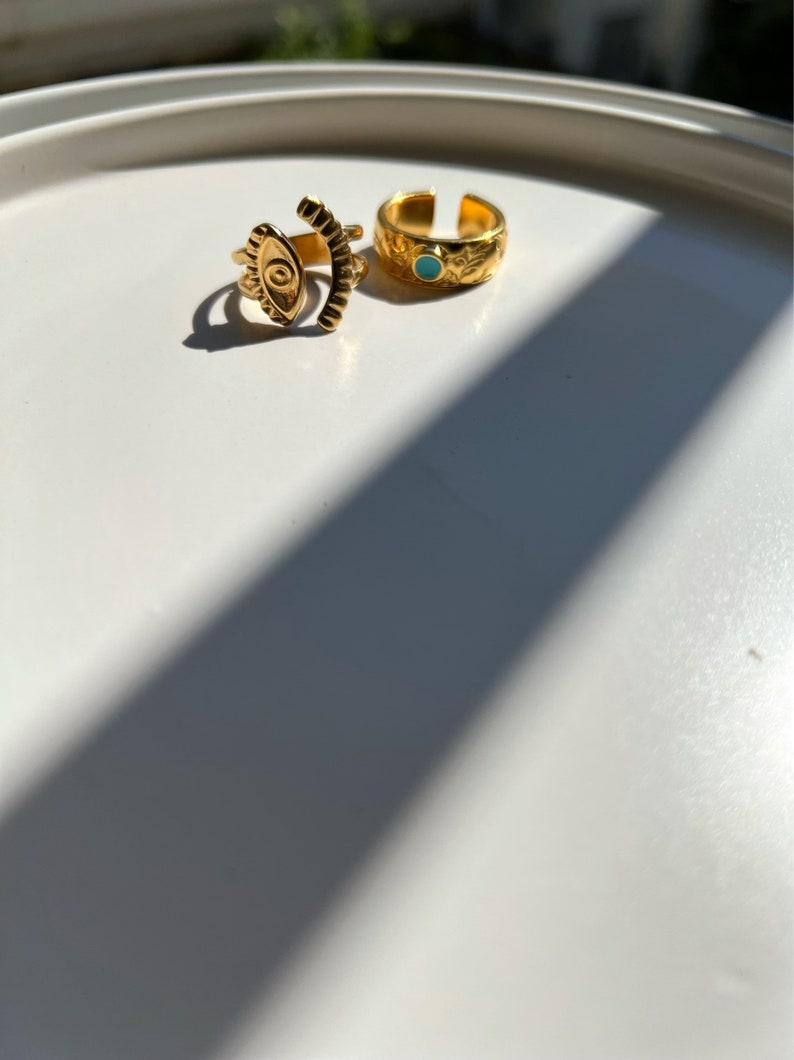 May include: Two gold rings on a white surface. One ring features an eye design and the other has a turquoise stone.
