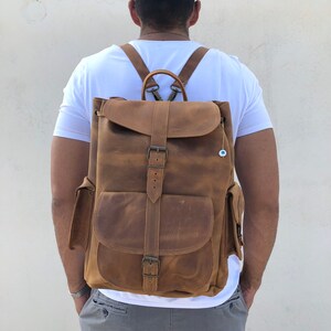 Puede incluir: Una mochila de cuero marrón con correas ajustables, un bolsillo frontal grande y un bolsillo más pequeño a cada lado. La mochila tiene un cierre de hebilla y un pequeño amuleto azul en forma de ojo.