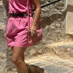 Op de afbeelding: Een roze, mouwloze jumpsuit met een zwarte riem en een roze leren schoudertas. De persoon staat op stenen trappen.