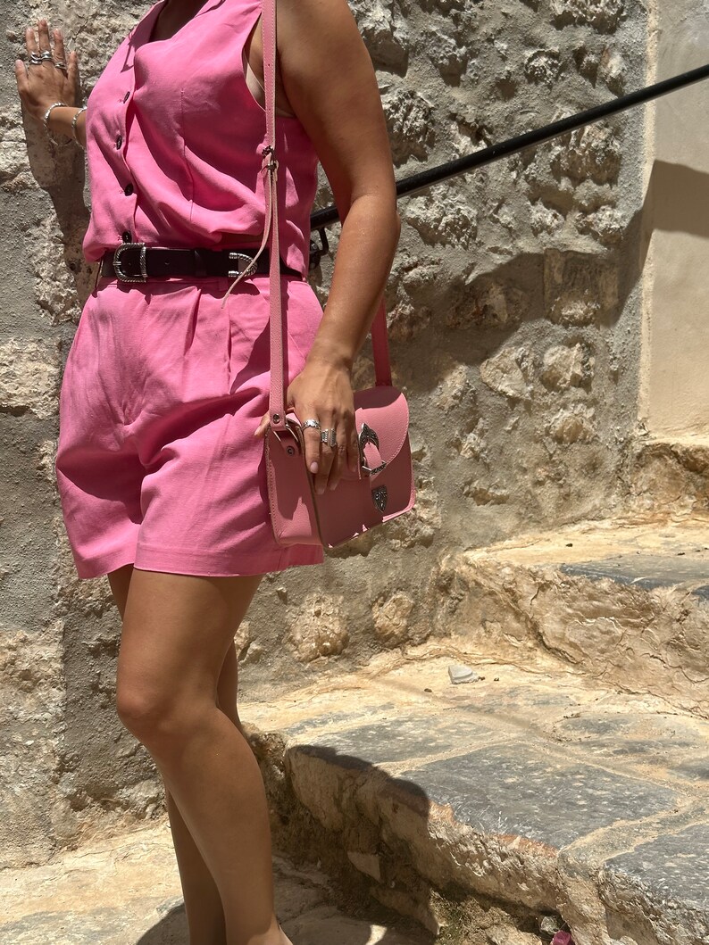 Op de afbeelding: Een roze, mouwloze jumpsuit met een zwarte riem en een roze leren schoudertas. De persoon staat op stenen trappen.