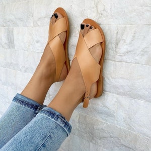 Puede incluir: Sandalias de cuero color canela con tiras cruzadas y hebilla en el tobillo. Las sandalias se llevan puestas en los pies de una persona con pantalones vaqueros azules.