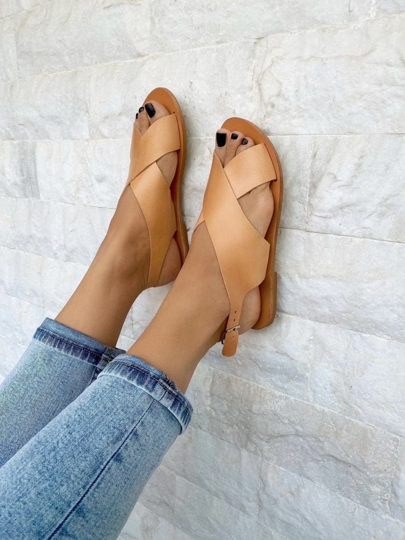Puede incluir: Sandalias de cuero color canela con tiras cruzadas y hebilla en el tobillo. Las sandalias se llevan puestas en los pies de una persona con pantalones vaqueros azules.