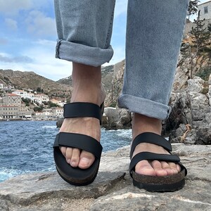May include: Black sandals with two adjustable straps and a cork footbed. The sandals are worn with light blue jeans. The image is taken outdoors with a coastal background.