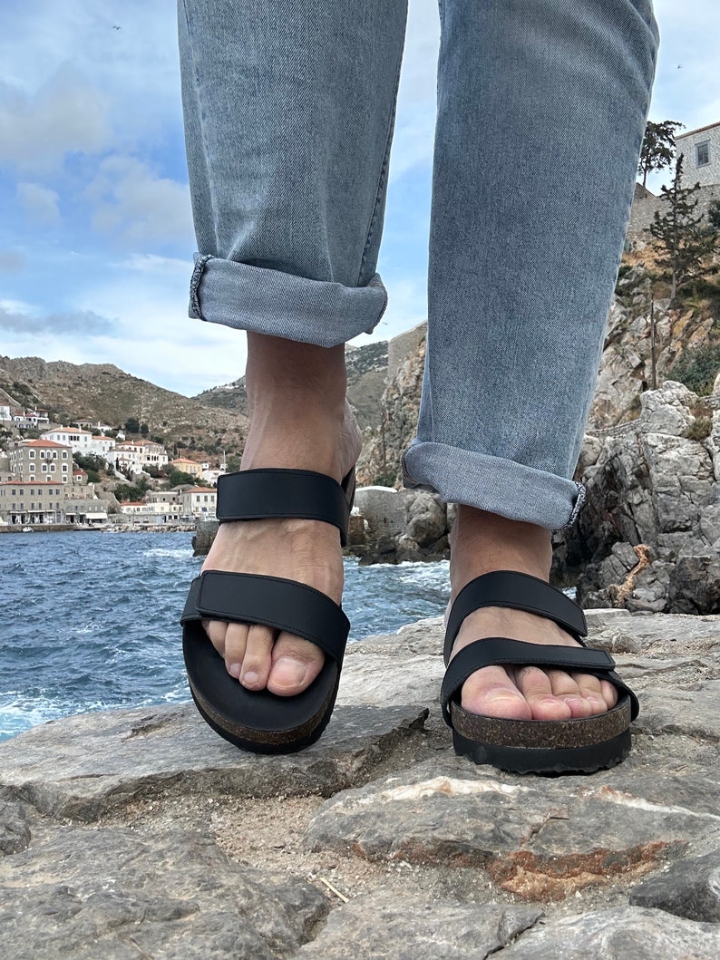 May include: Black sandals with two adjustable straps and a cork footbed. The sandals are worn with light blue jeans. The image is taken outdoors with a coastal background.