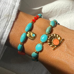 May include: Two turquoise beaded bracelets with gold accents and charms. One bracelet features a gold charm with an eye design, and the other has a gold seashell charm with an orange center. The bracelets are on a wrist.