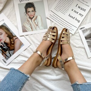 Op de afbeelding: Een paar gouden sandalen met een slangenhuidpatroon en een bandje om de enkel. De sandalen liggen op een wit oppervlak met tijdschriften op de achtergrond.