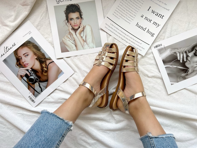 Op de afbeelding: Een paar gouden sandalen met een slangenhuidpatroon en een bandje om de enkel. De sandalen liggen op een wit oppervlak met tijdschriften op de achtergrond.