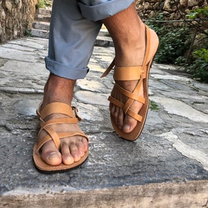 May include: Tan leather sandals with multiple straps crossing over the foot. The sandals have a simple design and are worn by a person standing on a stone path.