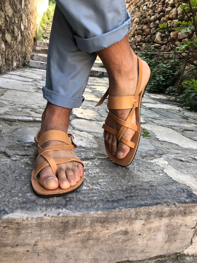 May include: Tan leather sandals with multiple straps crossing over the foot. The sandals have a simple design and are worn by a person standing on a stone path.