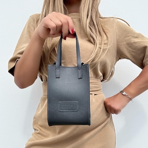 Könnte beinhalten: Eine kleine, dunkelgraue Lederhandtasche mit rechteckiger Form und zwei kurzen Henkeln. Die Tasche hat eine strukturierte Oberfläche und ein rechteckiges Etikett mit dem Text "CHRISTINA CHRISTI".