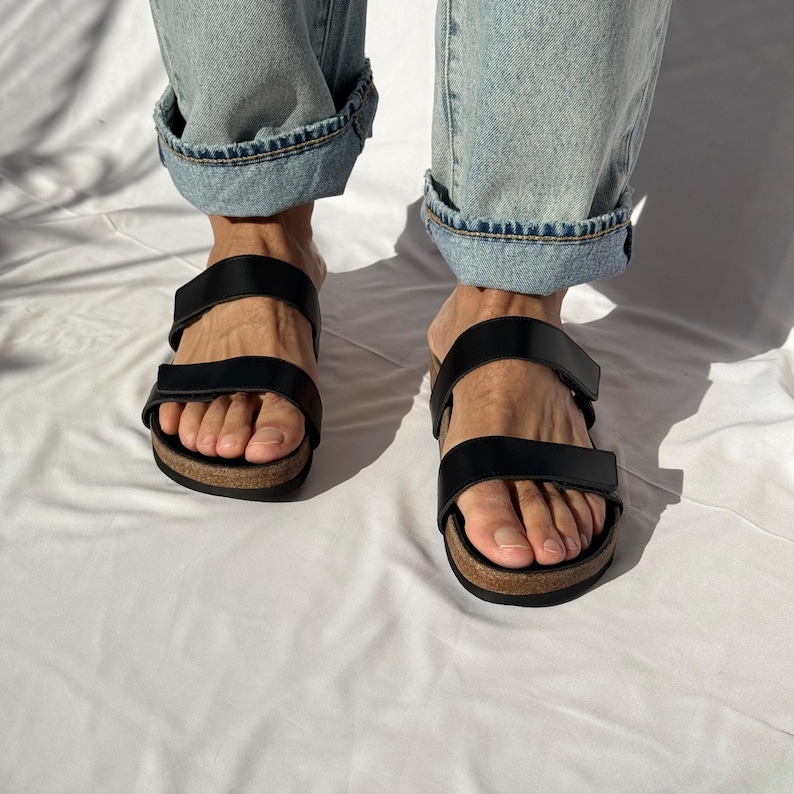 May include: Black leather sandals with two adjustable straps and a cork footbed. The sandals are worn with light blue denim jeans. The sandals are a casual footwear choice.