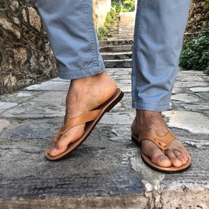 Pode incluir: Chinelos de couro castanho claro com sola castanha escura. As sand&aacute;lias s&atilde;o usadas com cal&ccedil;as azuis claras. A imagem &eacute; tirada ao ar livre em um caminho de pedra.