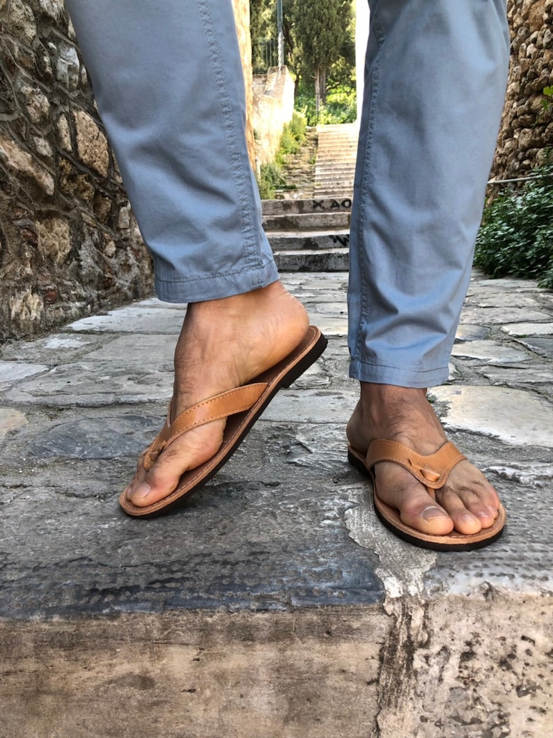 Pode incluir: Chinelos de couro castanho claro com sola castanha escura. As sand&aacute;lias s&atilde;o usadas com cal&ccedil;as azuis claras. A imagem &eacute; tirada ao ar livre em um caminho de pedra.